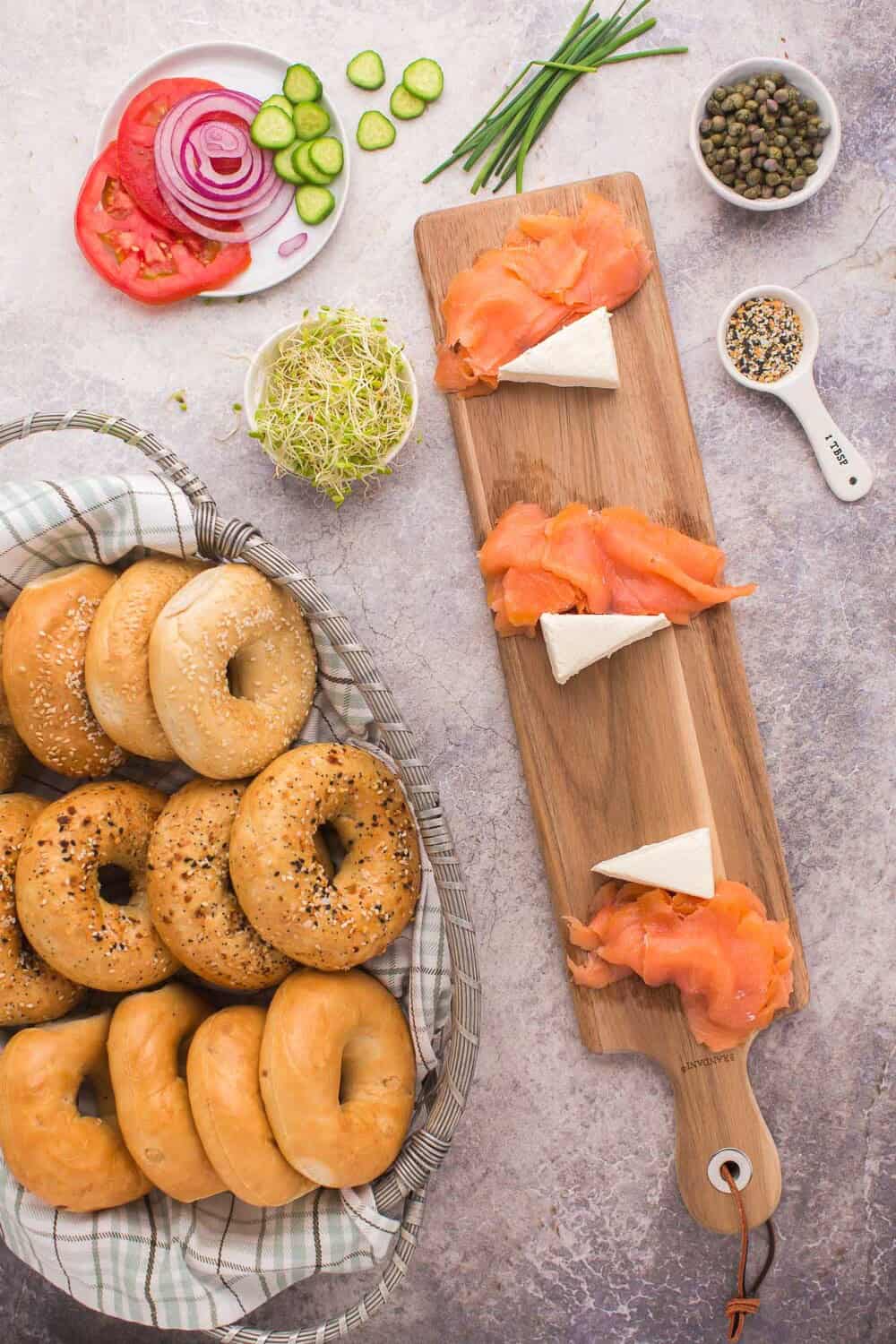A basket of assorted bagels beside a wooden board with smoked salmon and cheese slices. Surrounding items include sliced tomatoes, cucumbers, onion, capers, alfalfa sprouts, and seasoning.