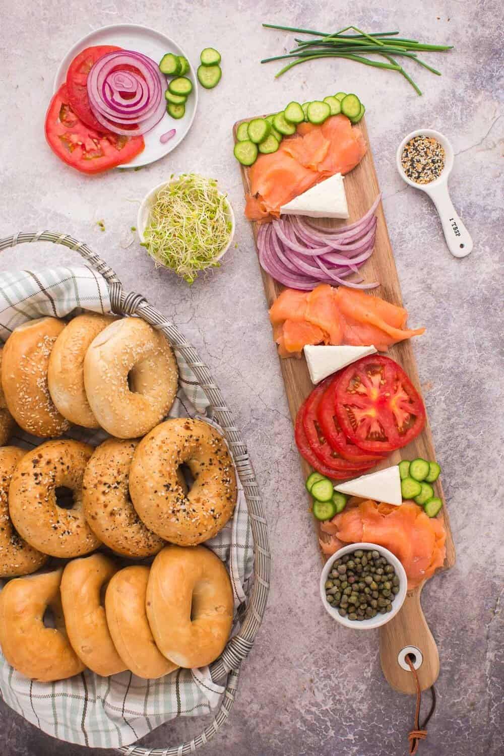 Bagels in a basket beside a board with smoked salmon, tomato slices, red onion, cucumber, cheese, and sprouts, garnished with seeds and capers.