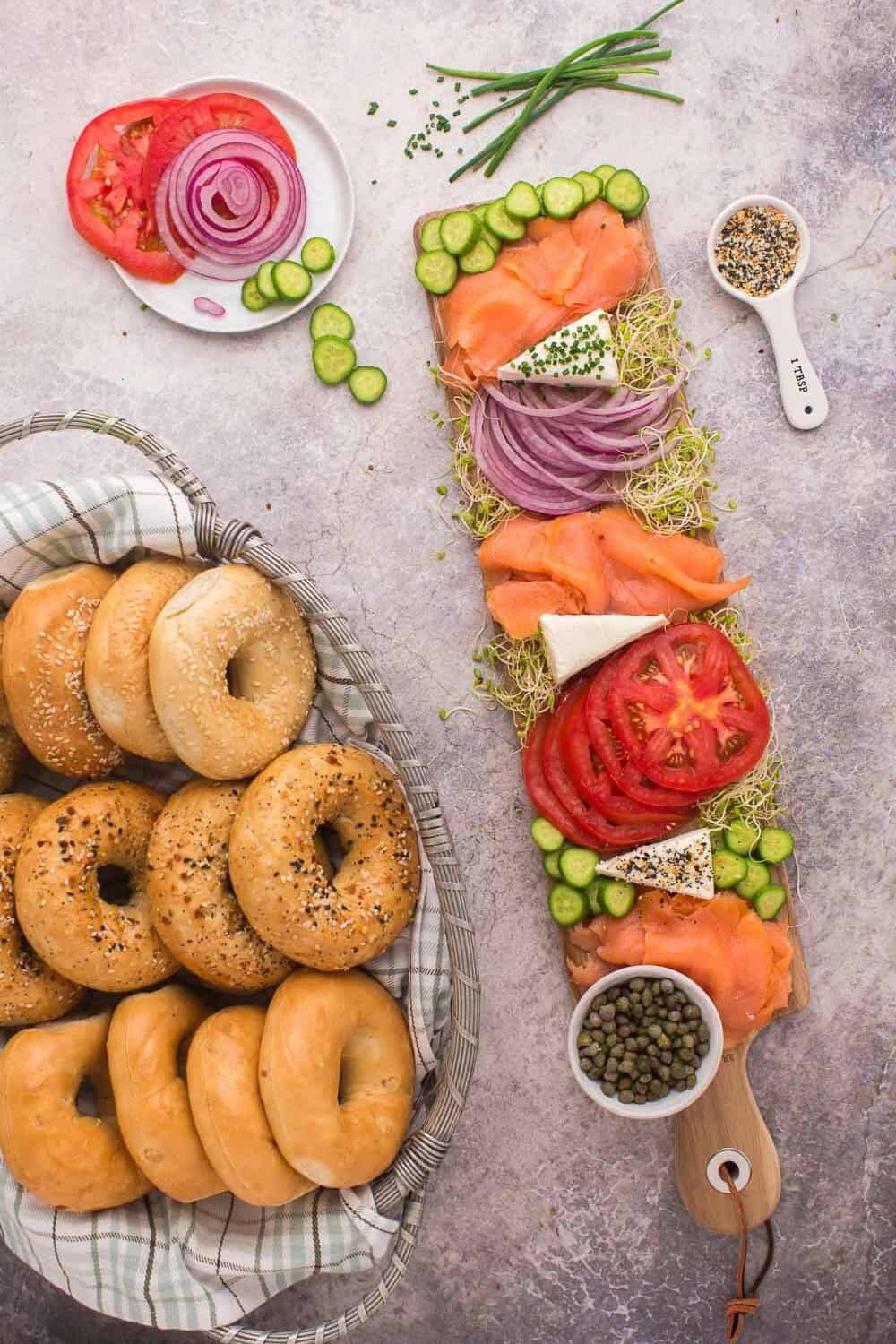 Bagels in a basket beside a board with smoked salmon, sliced tomatoes, onions, cucumbers, cheese, and capers. A small plate holds more sliced tomatoes and onions.