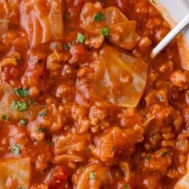 A close-up of a hearty soup with chunks of meat, tomatoes, cabbage, and herbs. A spoon is partially visible.