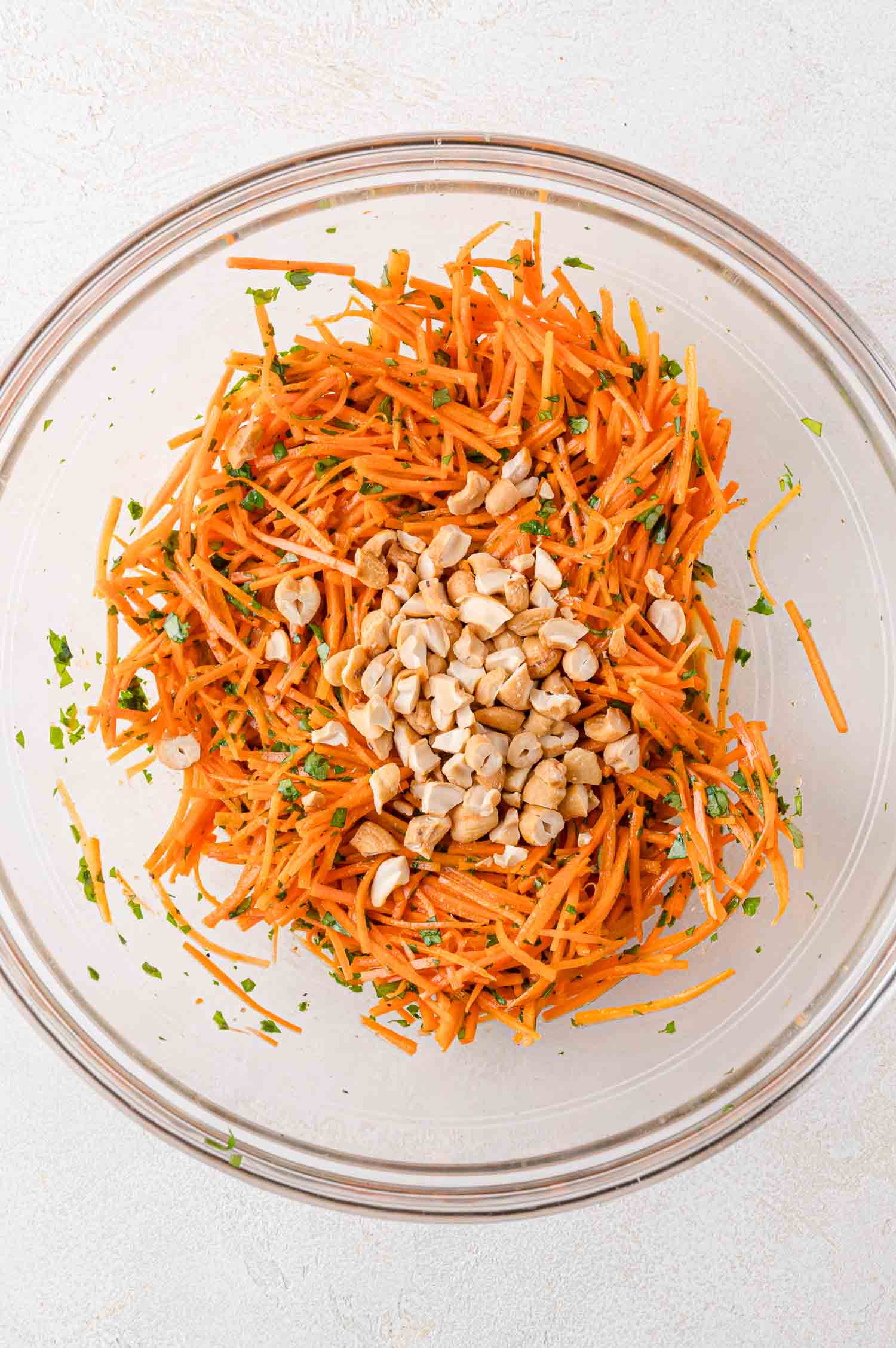 A glass bowl containing shredded carrots mixed with herbs, topped with chopped cashews on a light surface.