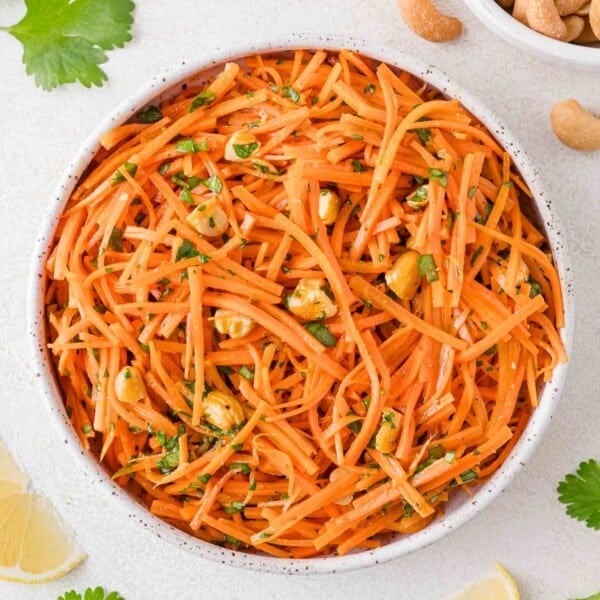 A bowl of carrot salad with shredded carrots, herbs, and cashew nuts on a light textured surface. Lemon slices and cilantro leaves are nearby.