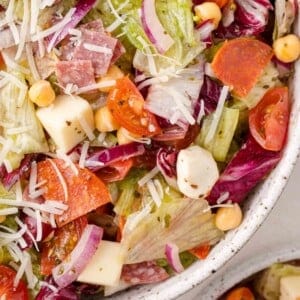 Close-up of a colorful salad featuring lettuce, cherry tomatoes, red onion, chickpeas, cheese cubes, sliced pepperoni, and shredded cheese in a speckled bowl.
