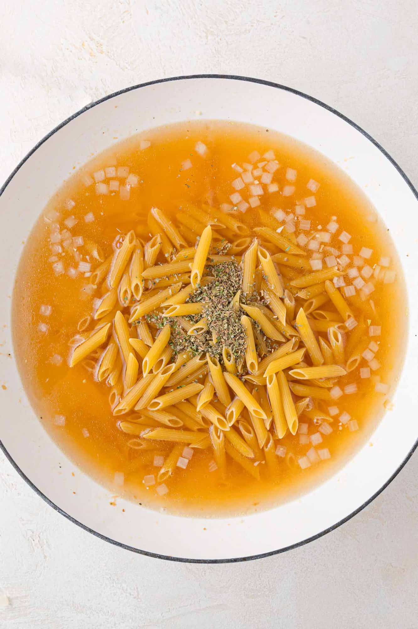 Uncooked penne pasta in broth with diced onions and herbs in a white pot.