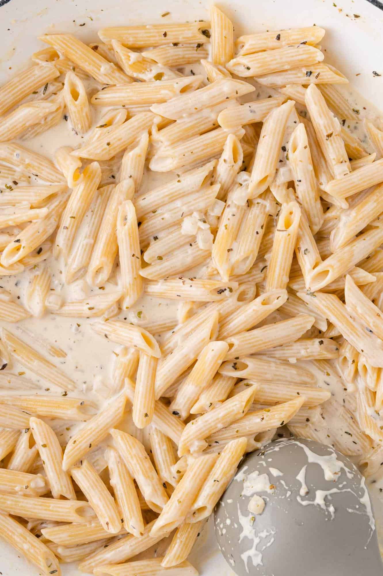Penne pasta in a creamy white sauce with herbs in a skillet, accompanied by a gray spatula.