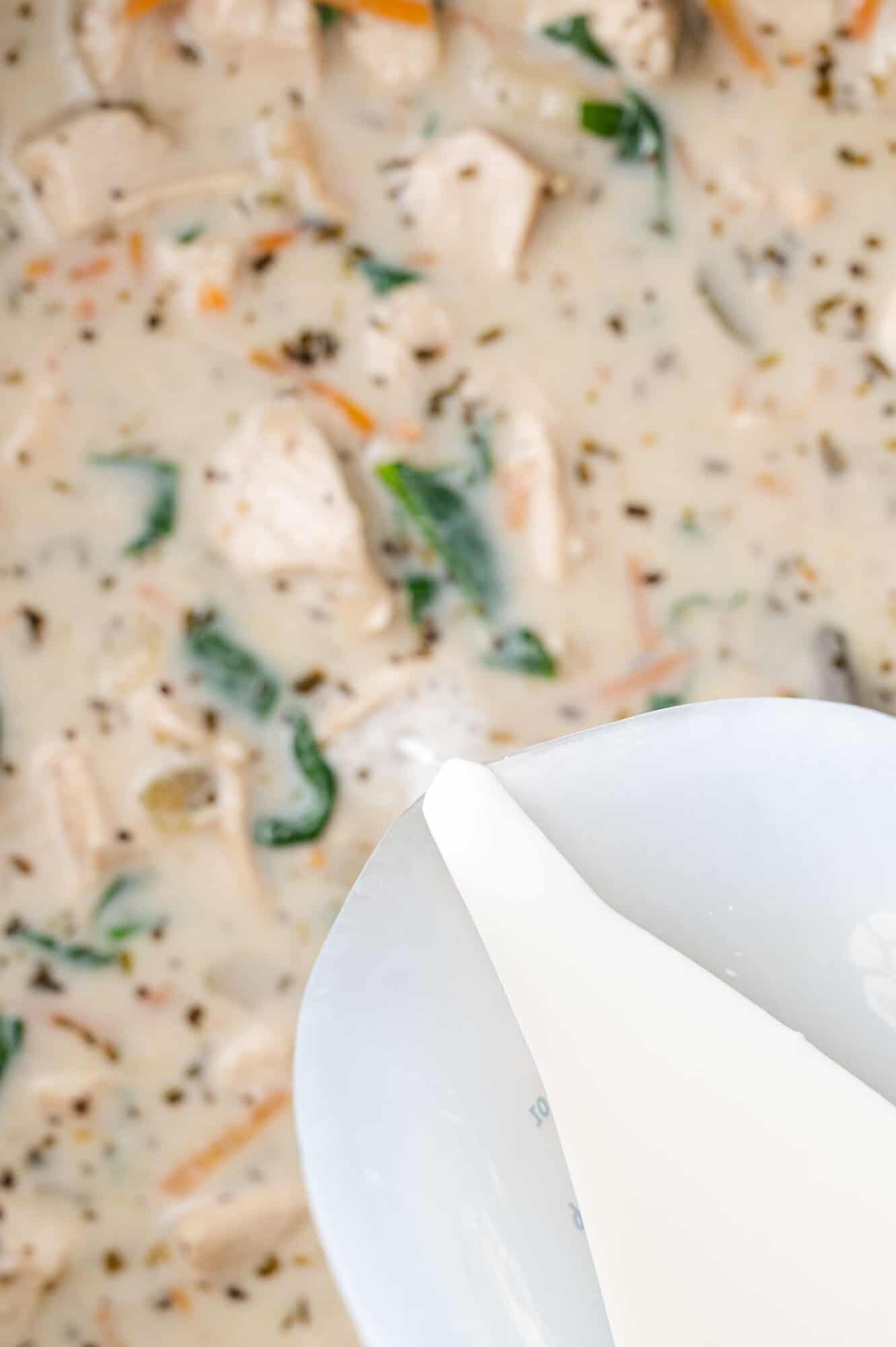 Pouring milk into a creamy soup with chunks of chicken, spinach, and herbs.