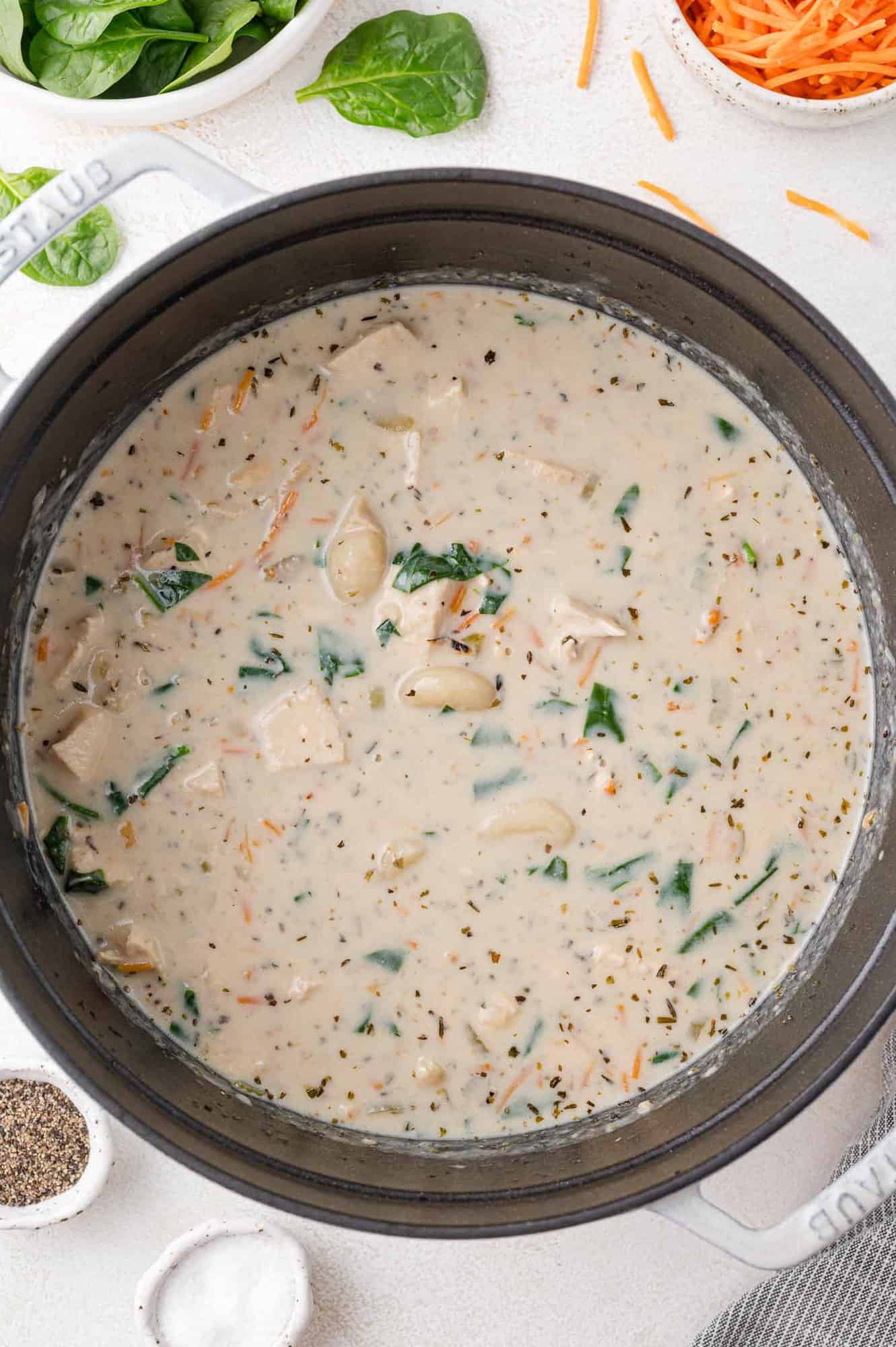 A pot of creamy chicken soup with pasta, spinach, and carrots on a white surface, surrounded by small bowls of salt and pepper.