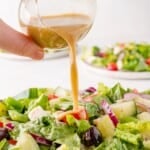 A hand pours dressing over a fresh Greek salad with lettuce, cucumbers, tomatoes, red onions, and olives on a plate.