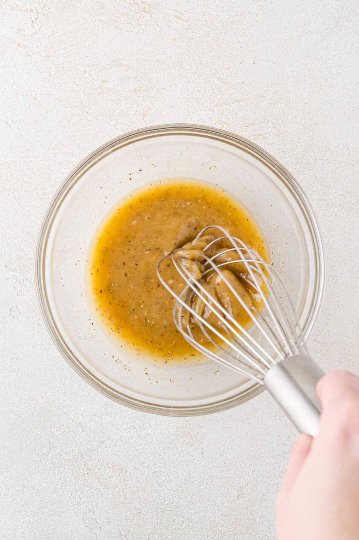 A hand whisking dressing in a clear glass bowl. The liquid is a light golden color with visible herbs and seasoning.