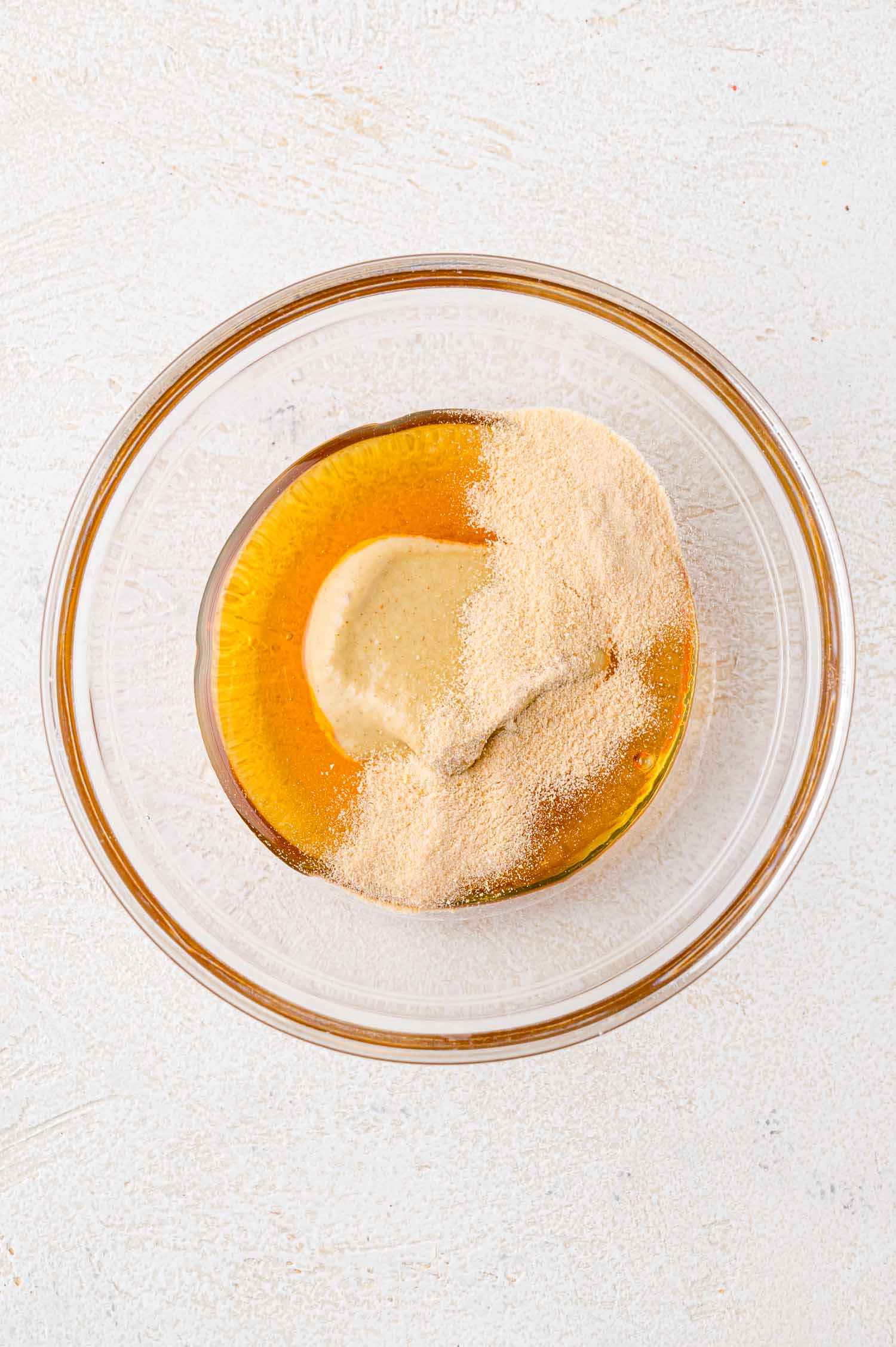 A glass bowl with liquid honey, mustard, and powder on a light textured surface.
