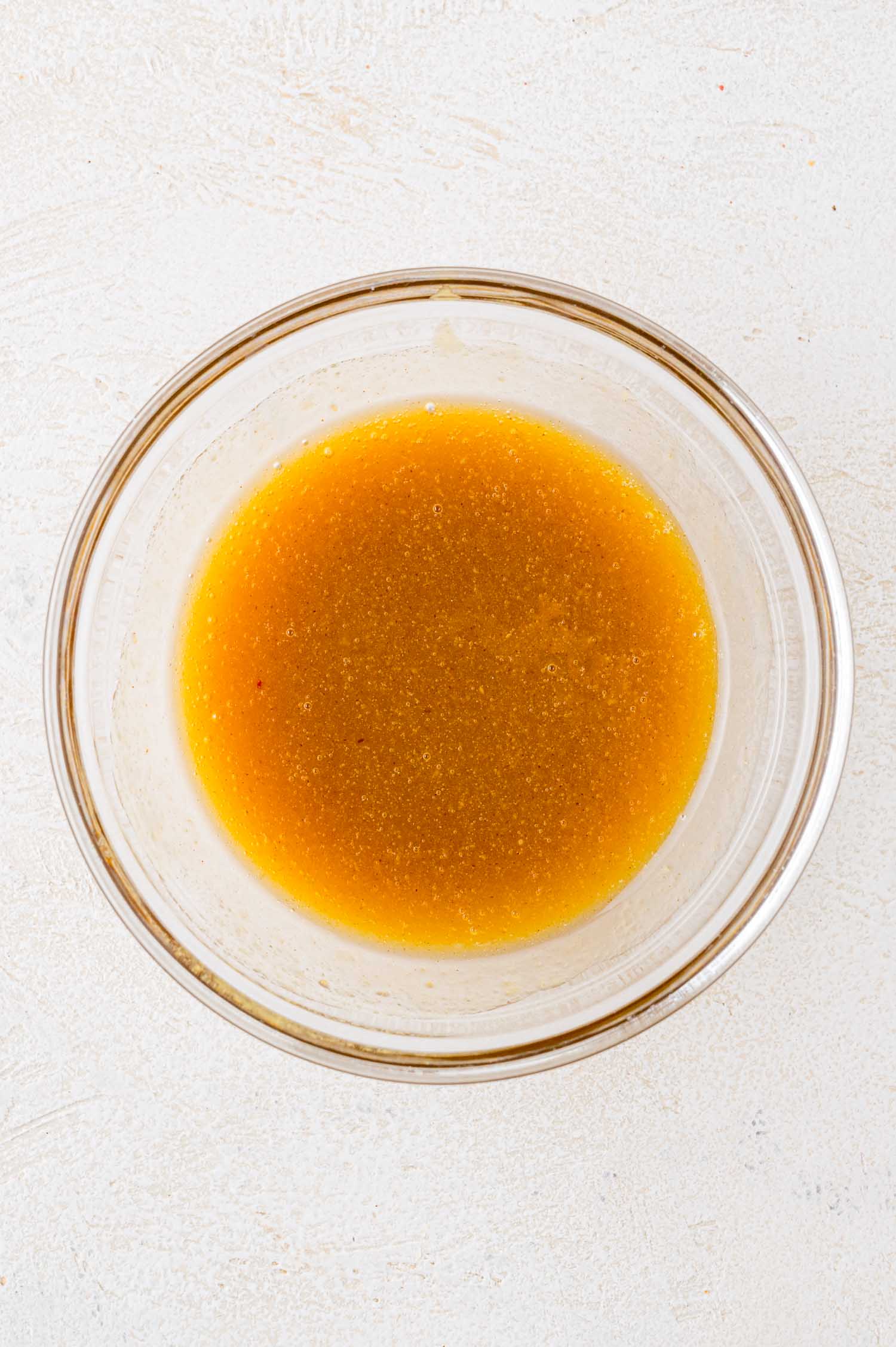 A glass bowl filled with a light brown liquid sauce on a white textured surface.