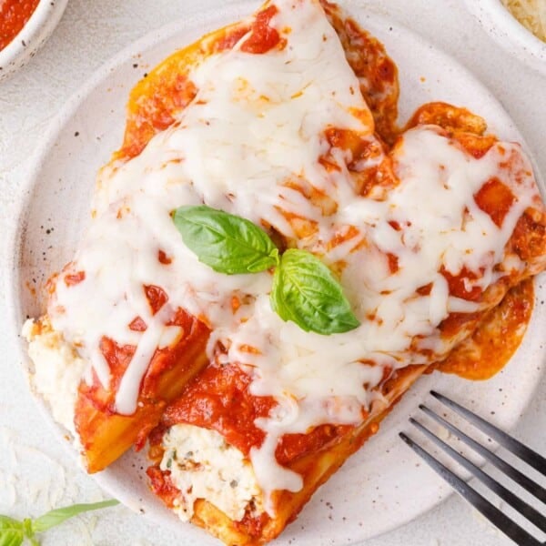 Plate of manicotti topped with tomato sauce, melted cheese, and basil leaves. Fork on the side.