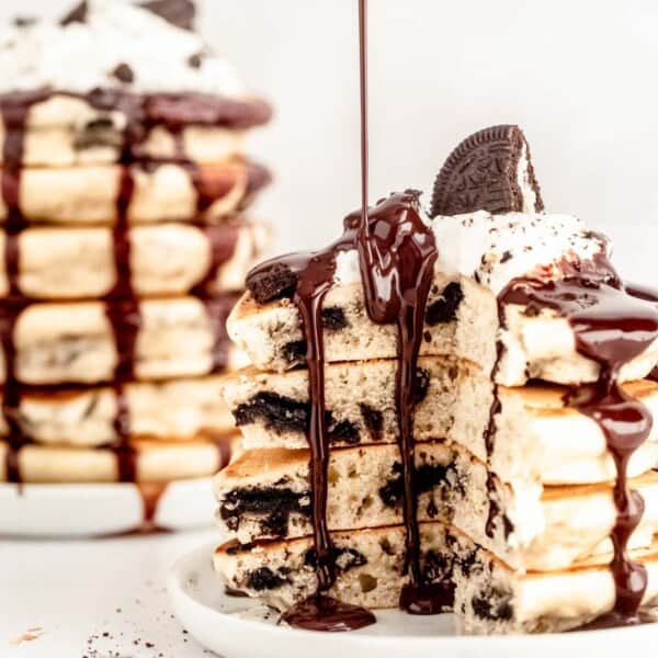 Stack of Oreo pancakes topped with whipped cream and an Oreo cookie, with chocolate sauce being poured over them. Another stack is visible in the background.
