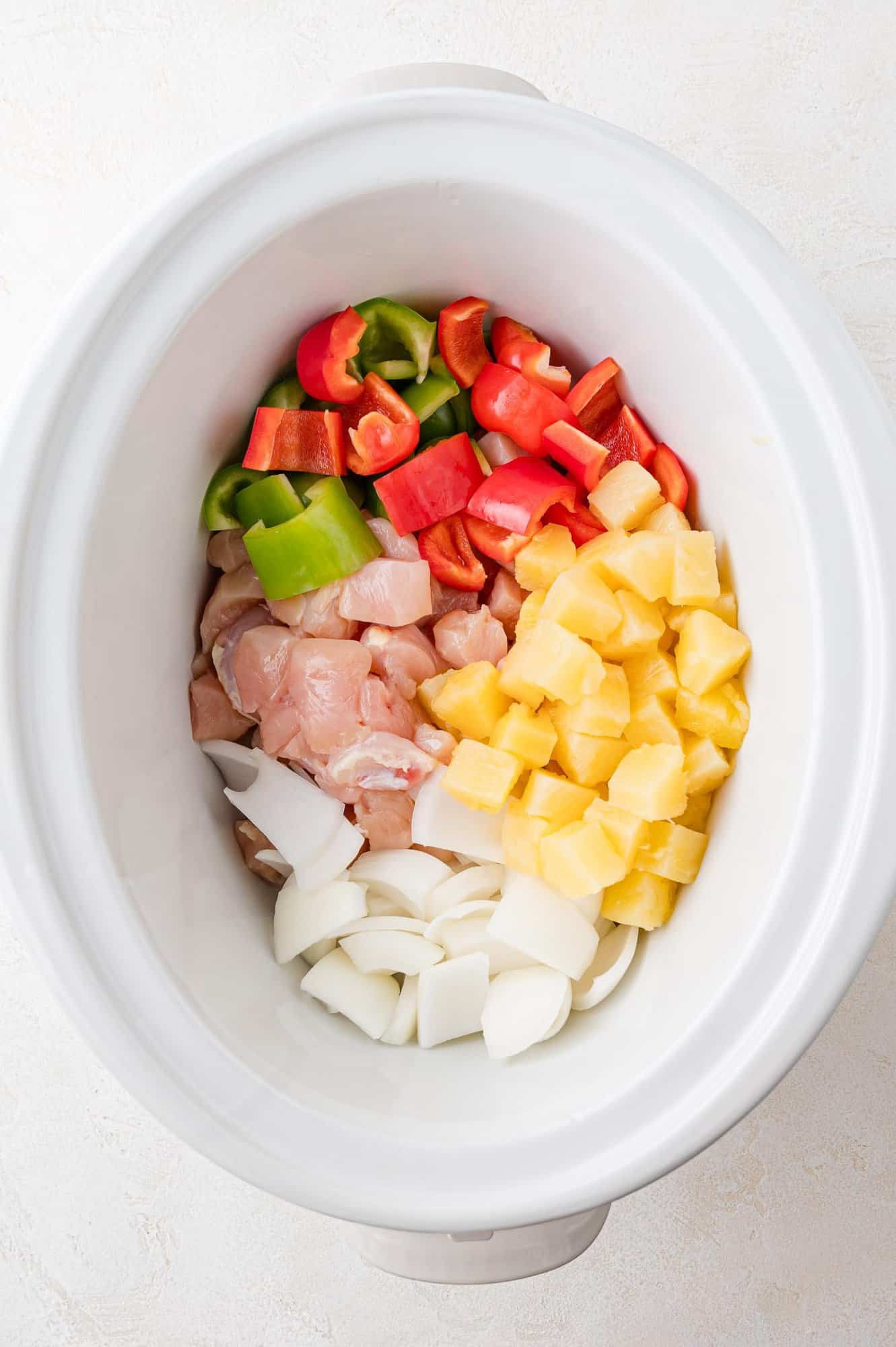 Chopped chicken, red and green bell peppers, pineapple, and onions arranged in a white slow cooker.
