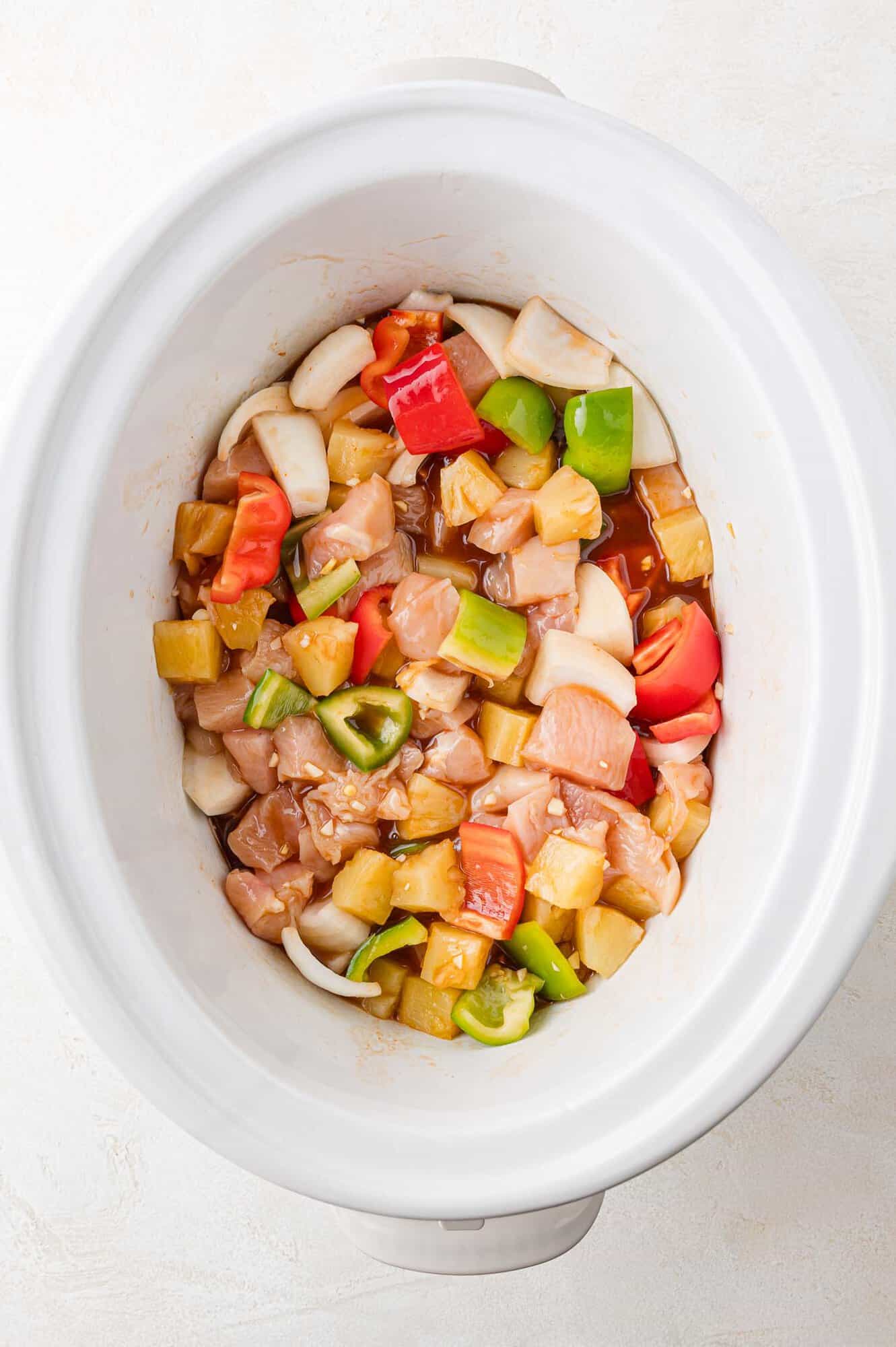Slow cooker filled with raw diced chicken, red and green bell peppers, pineapple chunks, and onions, ready for cooking.