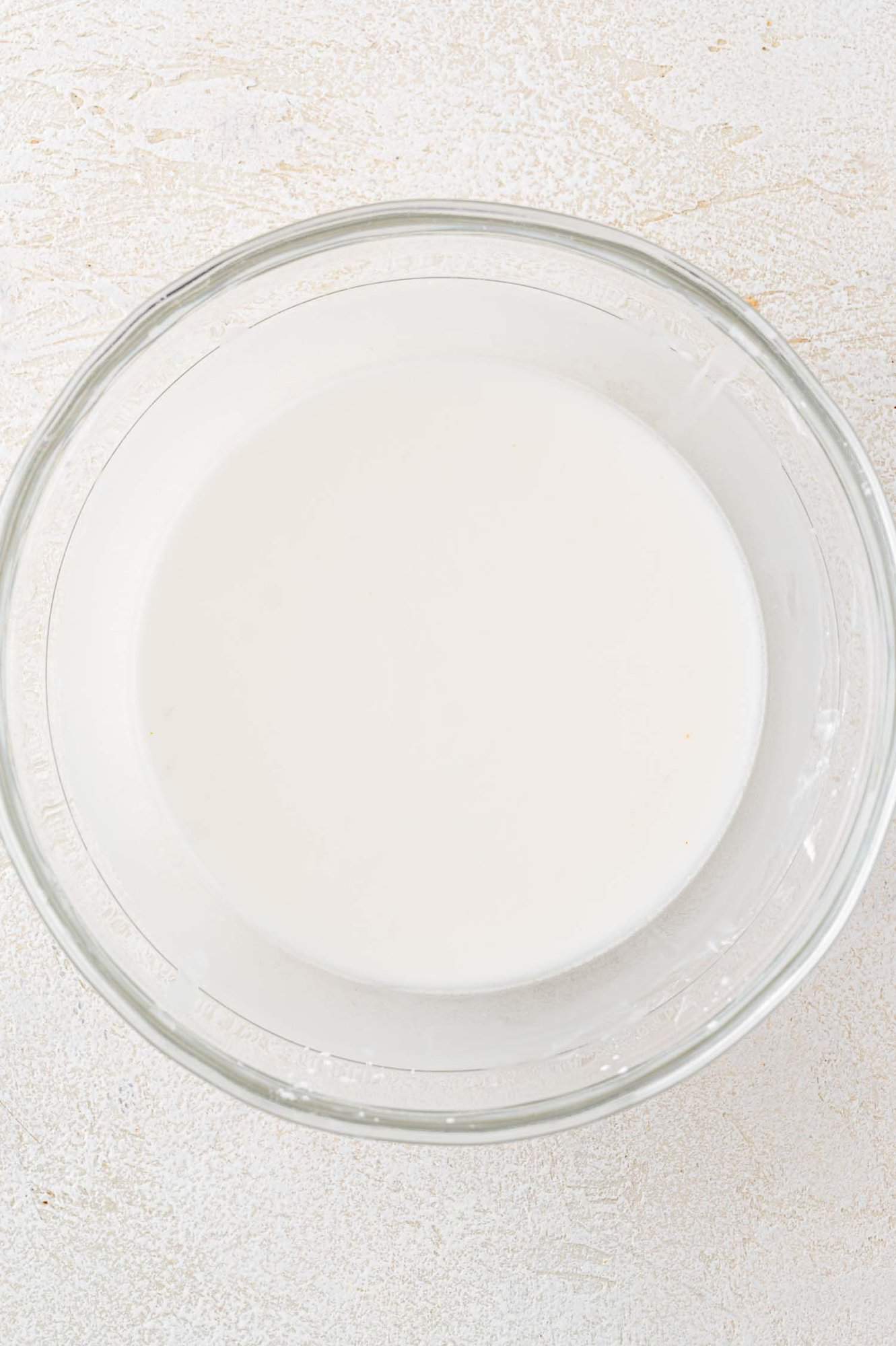 A clear glass bowl filled with white liquid on a textured white surface.