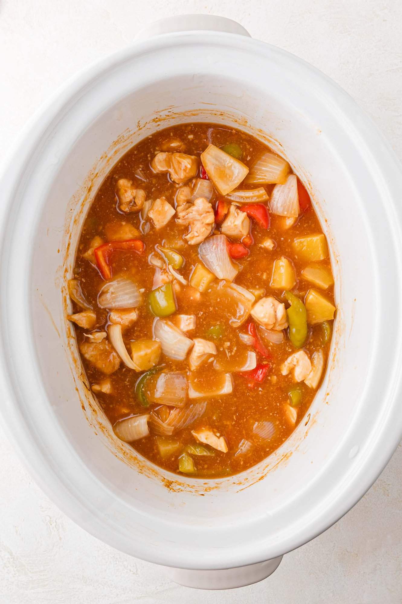 A white slow cooker containing Hawaiian chicken with chunks of chicken, onions, bell peppers, and a thick, reddish sauce.