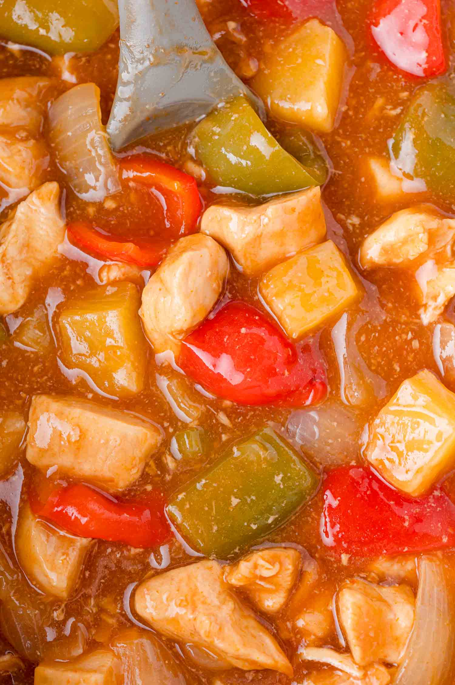 Close-up of Hawaiian chicken with chunks of chicken, red and green bell peppers, onions, and pineapple in a glossy sauce, being stirred with a gray spoon.