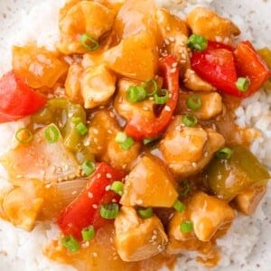 A plate of sweet Hawaiian chicken with diced chicken, pineapple, red and green bell peppers, and sauce over white rice, garnished with green onions and sesame seeds.