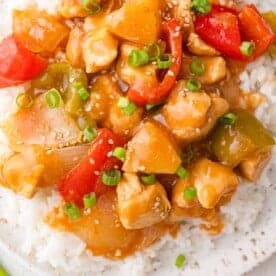A plate of sweet hawaiian chicken with bell peppers, onions, and pineapple on rice, garnished with sesame seeds and chopped green onions.