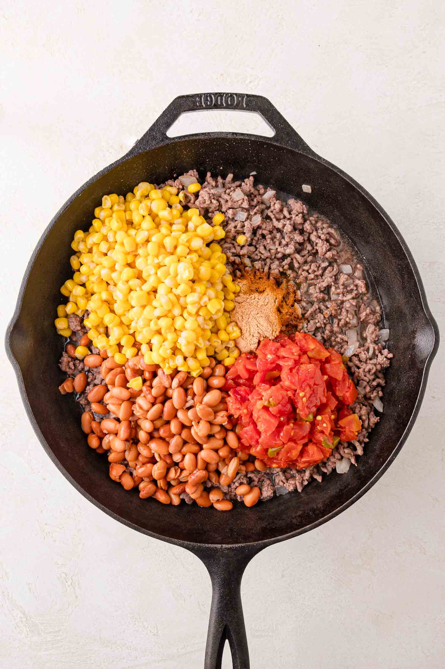 Skillet with cooked ground beef, corn, diced tomatoes, pinto beans, and seasoning.