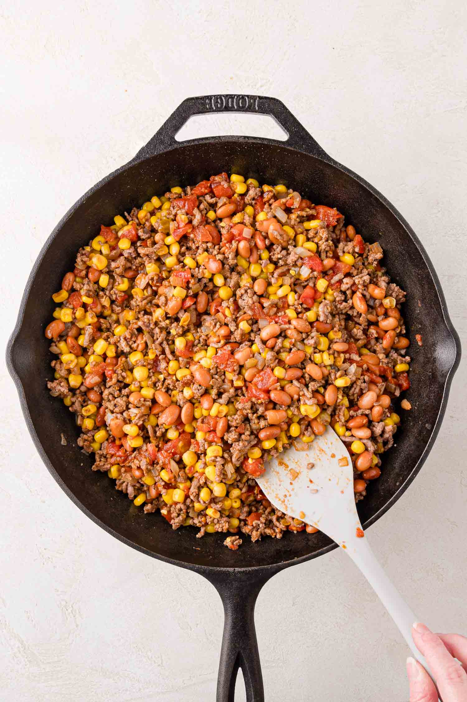 A skillet with a mixture of ground beef, pinto beans, corn, and diced tomatoes being stirred with a white spatula.