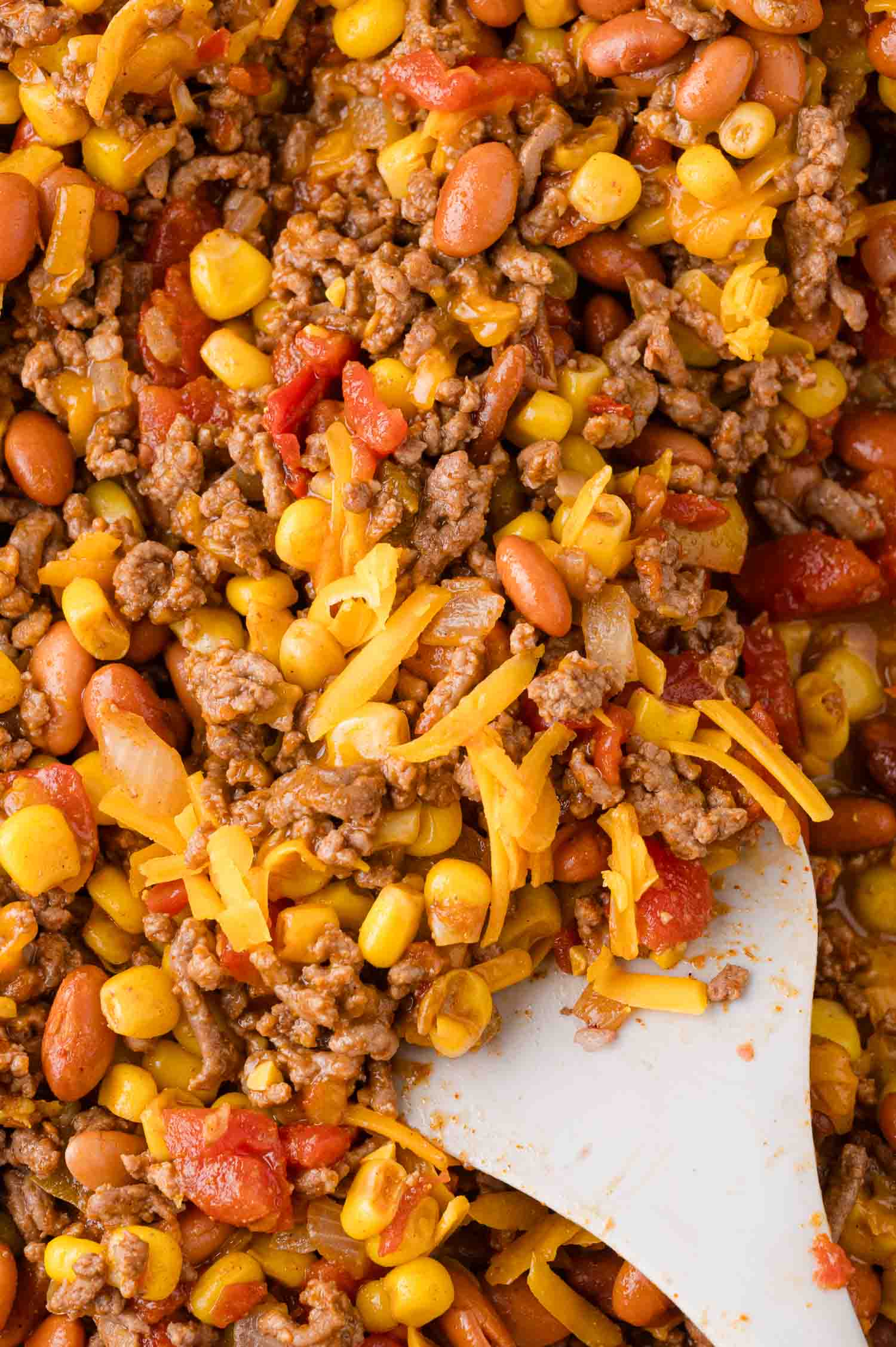 Close-up of a hearty mixture featuring ground beef, corn, kidney beans, tomatoes, and melted cheese, stirred with a white spatula.