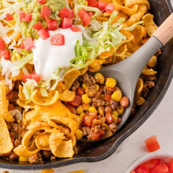 A skillet filled with taco ingredients, including ground beef, corn, beans, and topped with sour cream, guacamole, shredded lettuce, tomatoes, and corn chips.