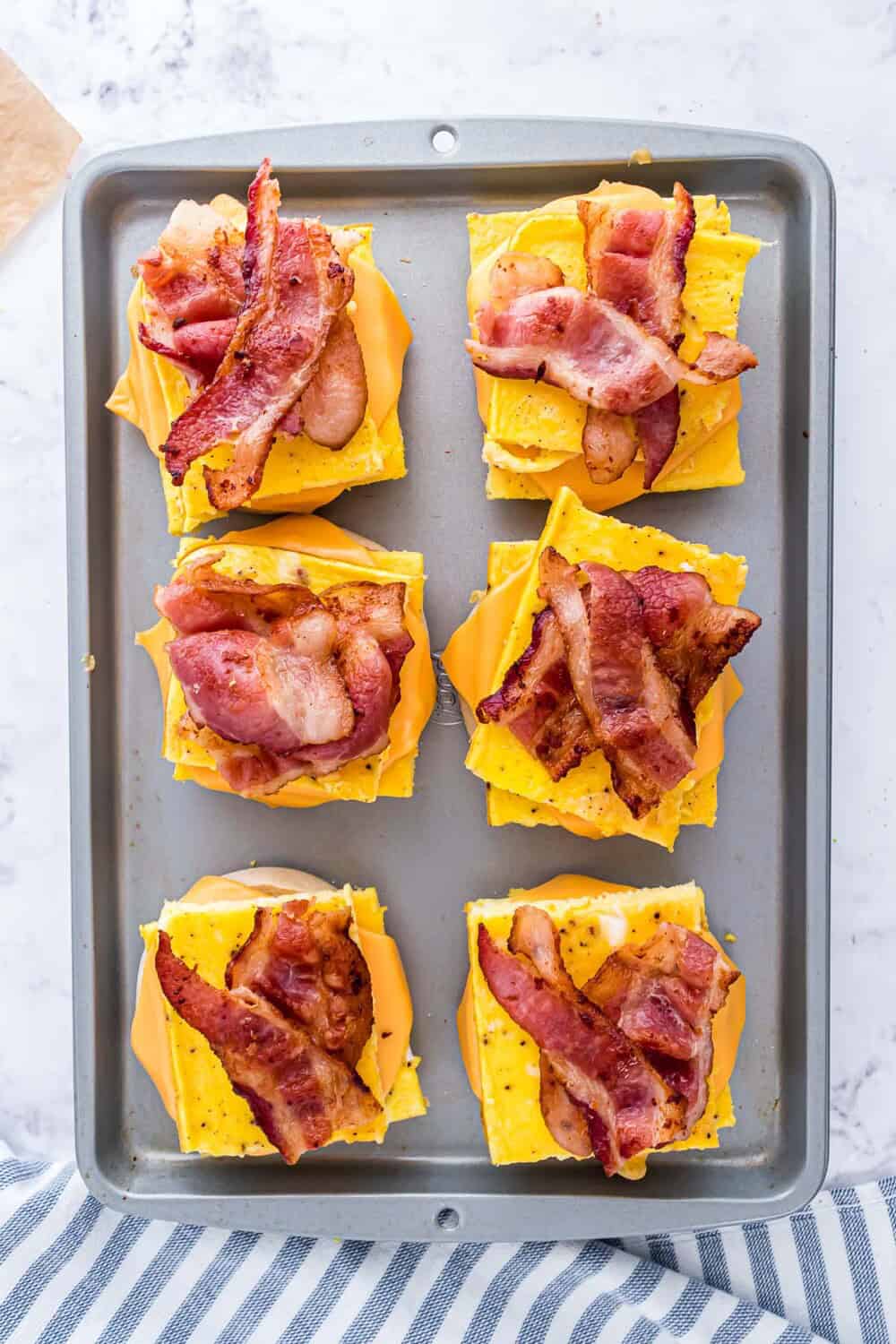 A baking tray with six breakfast sandwiches, each with scrambled eggs, melted cheese, and crispy bacon on a english muffin.