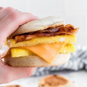 Hand holding a breakfast sandwich with bacon, eggs, and cheese between an English muffin.
