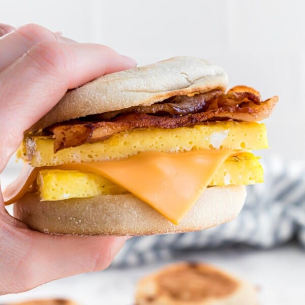 Hand holding a breakfast sandwich with bacon, eggs, and cheese between an English muffin.