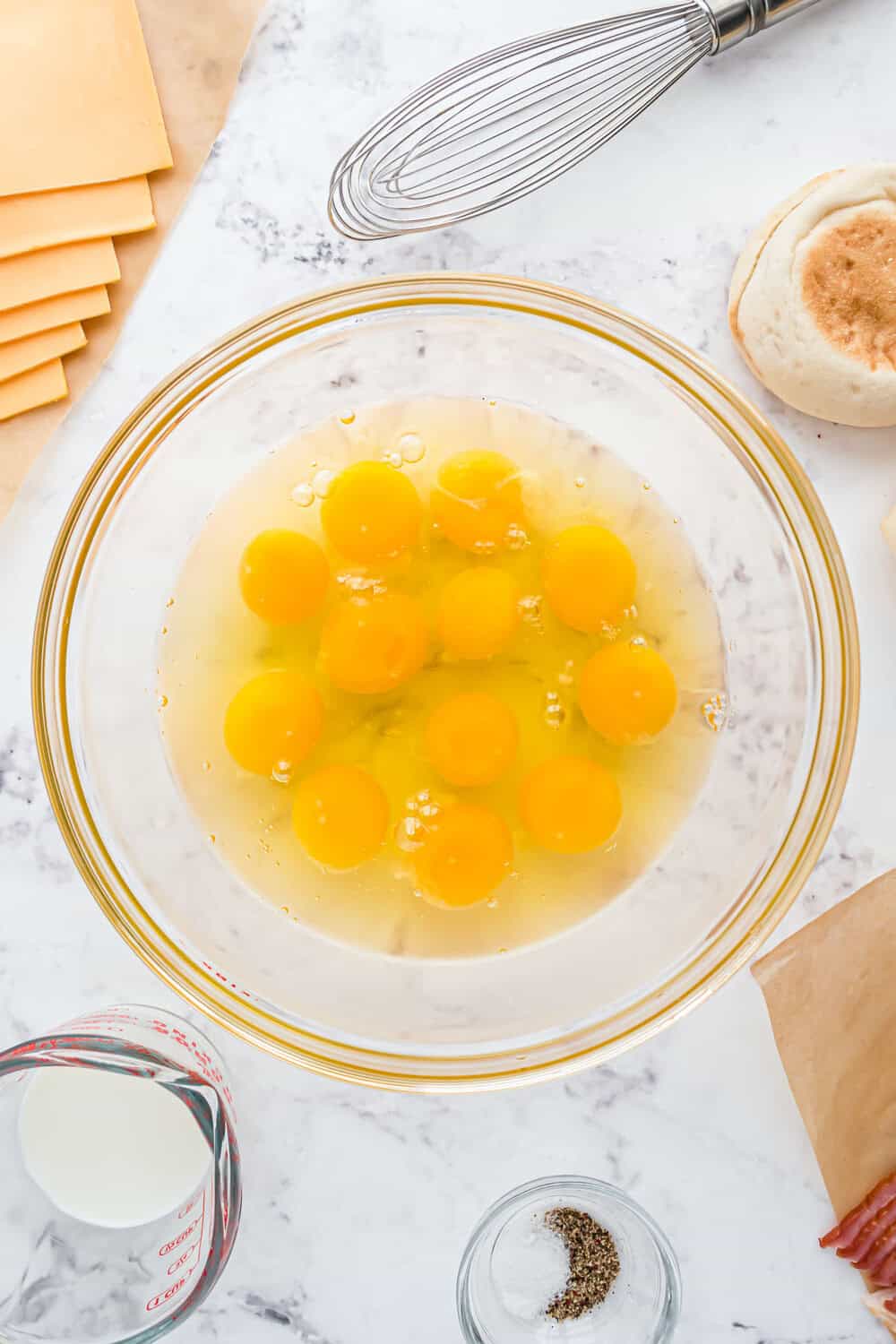 A glass bowl with a dozen raw eggs on a marble countertop, surrounded by a whisk, cheese slices, a measuring cup with milk, salt, pepper, and an English muffin.