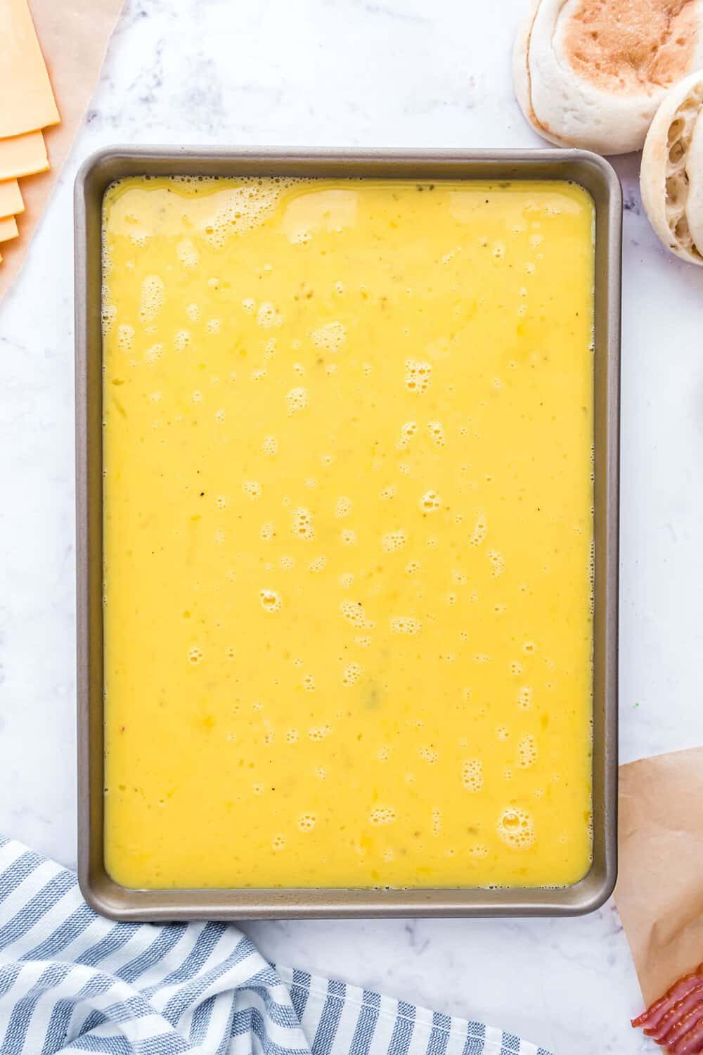 A rectangular baking dish filled with uncooked scrambled eggs. Cheese slices, English muffins, and a striped cloth are nearby on a marble countertop.