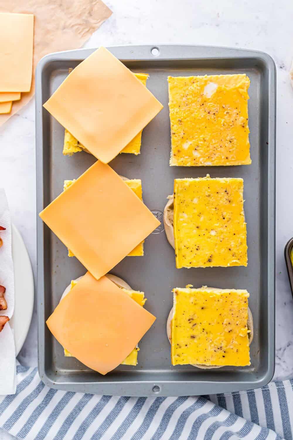 Cheese slices on top of scrambled egg squares on english muffins, placed on a baking tray.