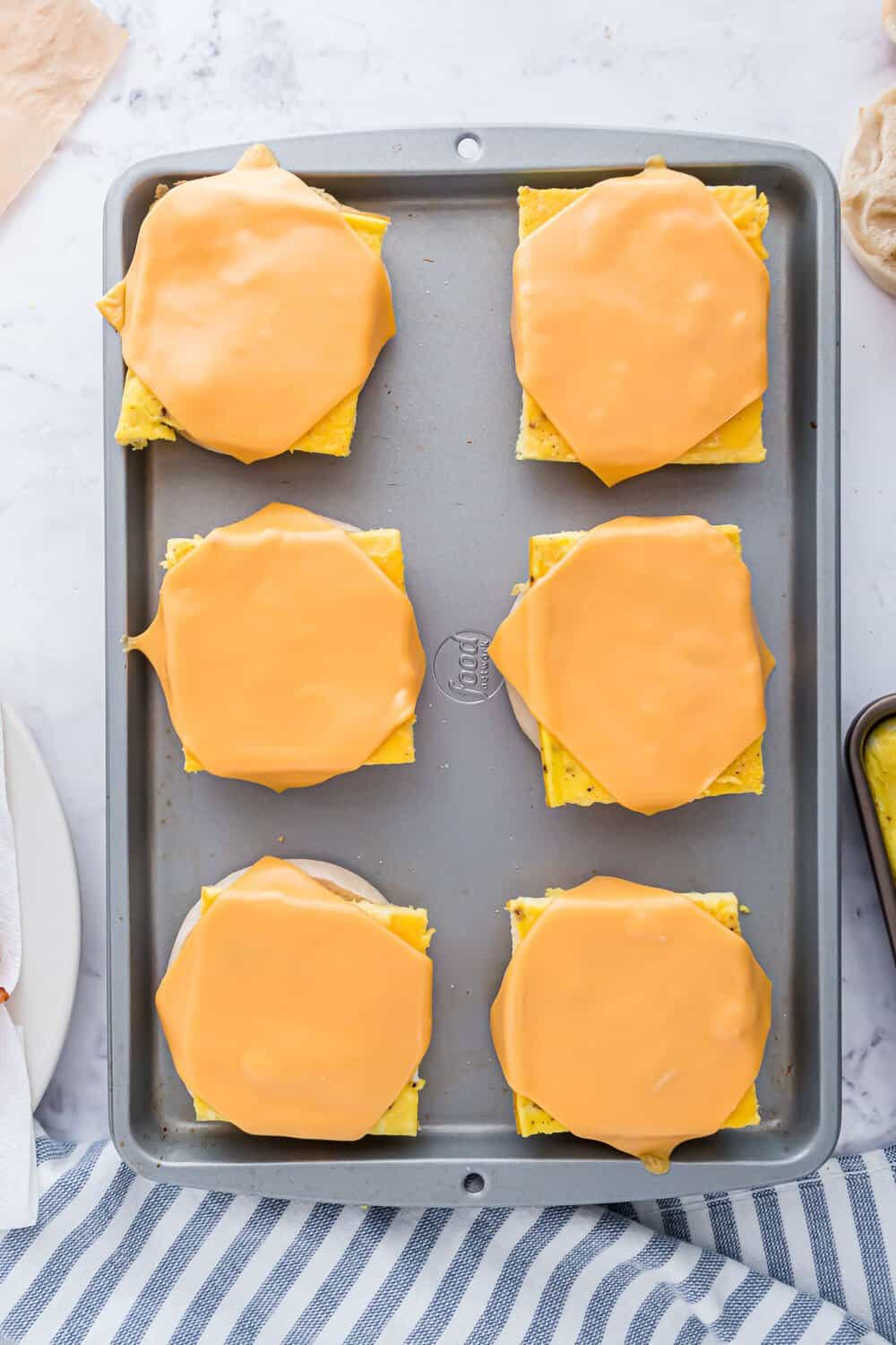 Baking sheet with six pieces of english muffins, each topped with scrambled eggs and a slice of melted cheese.