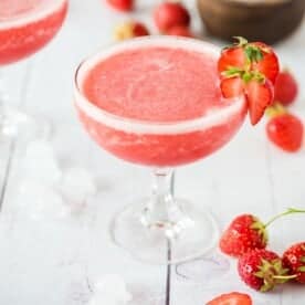 Frosé in a glass, garnished with a strawberry slice, surrounded by whole strawberries and ice cubes on a white surface.