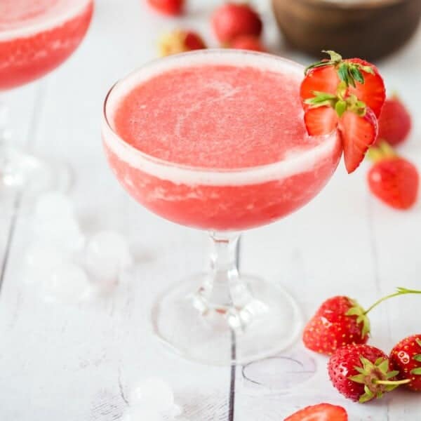 Frosé in a glass, garnished with a strawberry slice, surrounded by whole strawberries and ice cubes on a white surface.