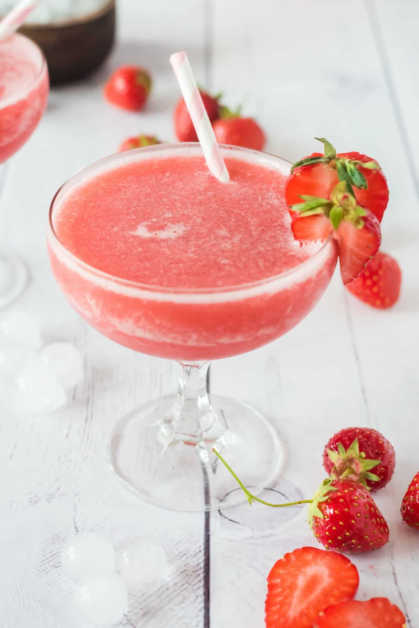A pink frozen strawberry rose cocktail in a glass with a straw and a fresh strawberry garnish.