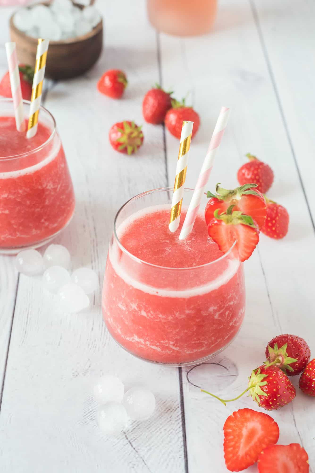 Two glasses of strawberry frose on a white wooden table, garnished with fresh strawberries and striped straws. 