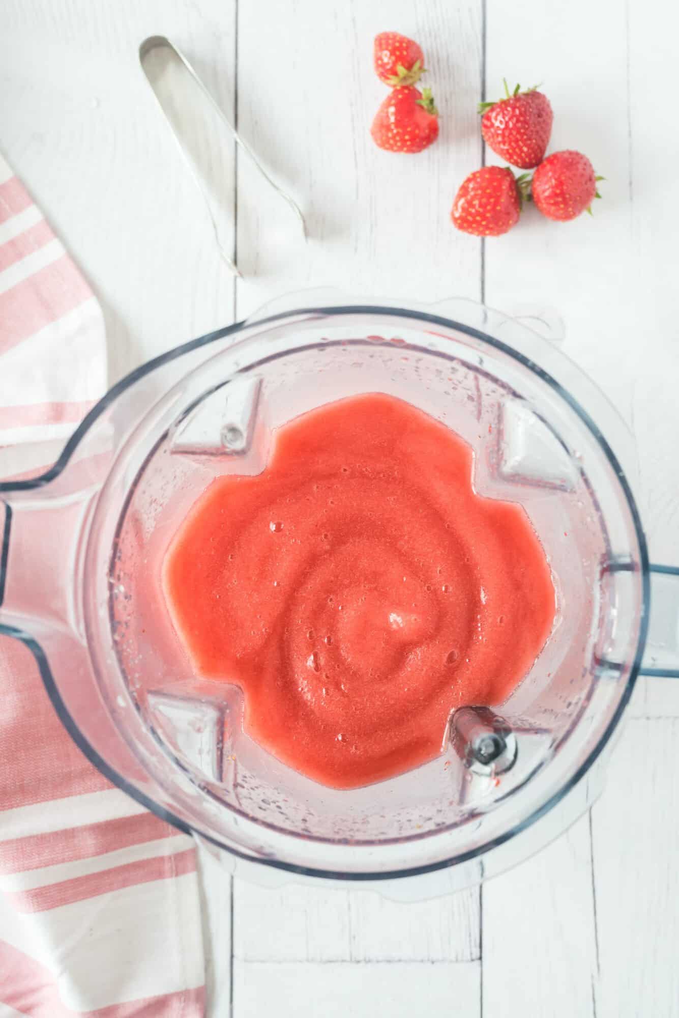 Blender with smooth frose on a white wooden surface, next to several whole strawberries and a pink-striped cloth.