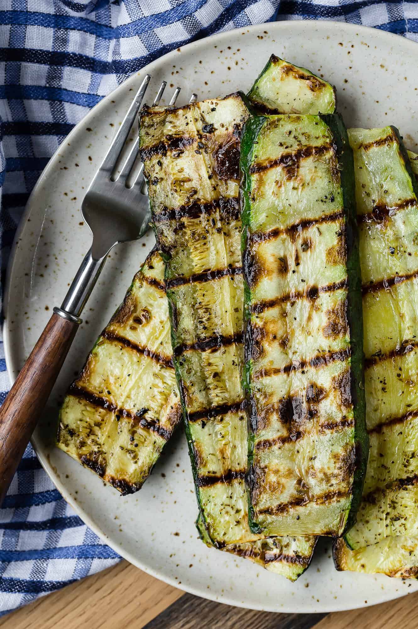 Grilled zucchini slices on a plate with a fork, set on a blue checkered cloth.