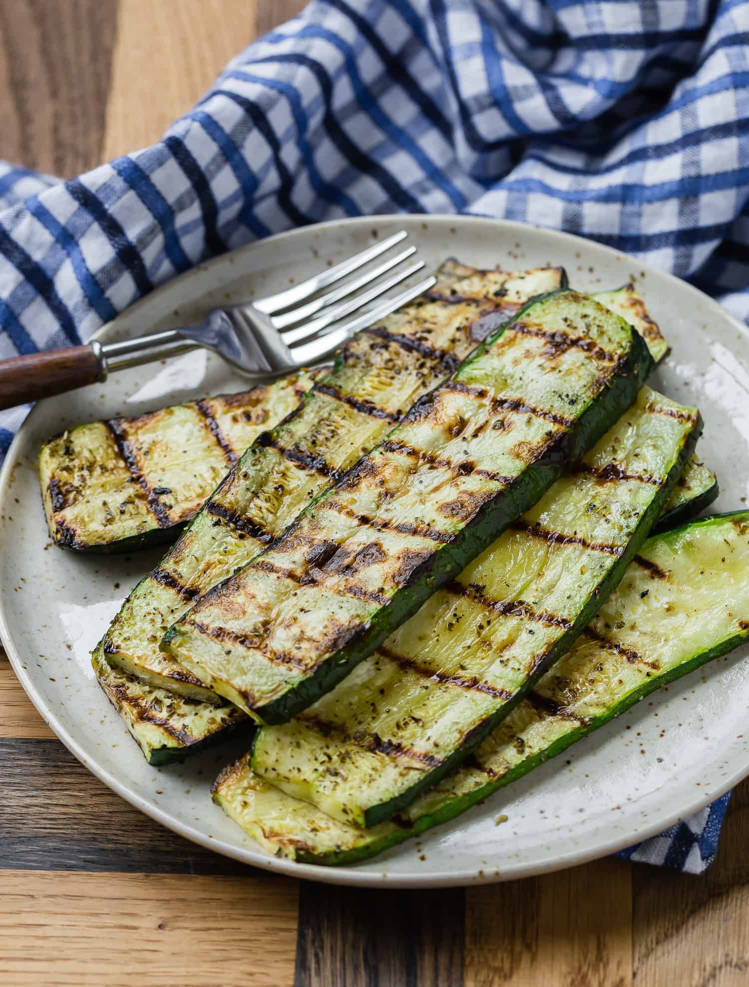 Grilled zucchini slices on a plate with a fork and a blue checkered cloth in the background.