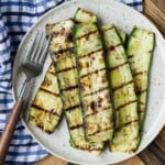 Grilled zucchini slices on a speckled plate beside a fork, with a blue and white checkered cloth underneath.