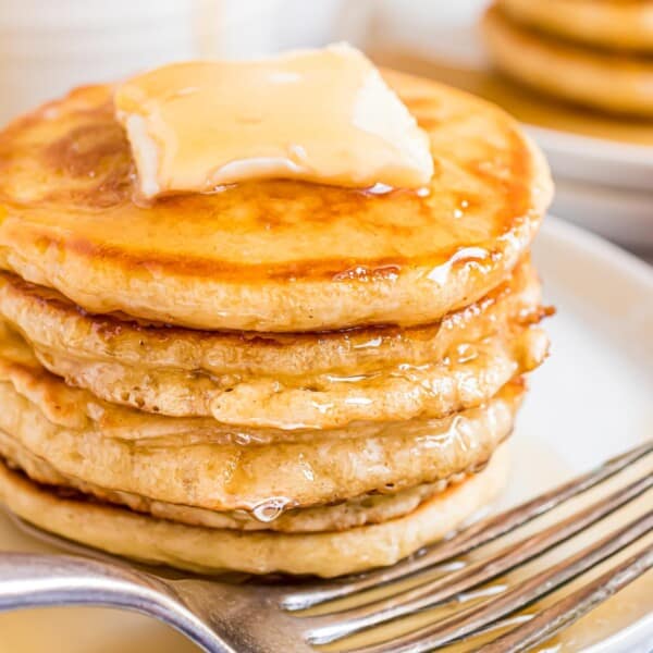 A stack of pancakes topped with a melting butter pat and syrup sits on a white plate, with a fork beside it.