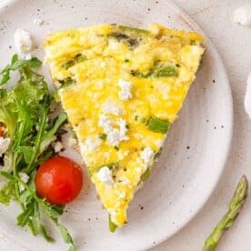 A slice of vegetable frittata with asparagus and goat cheese on a plate, accompanied by mixed greens and a cherry tomato.
