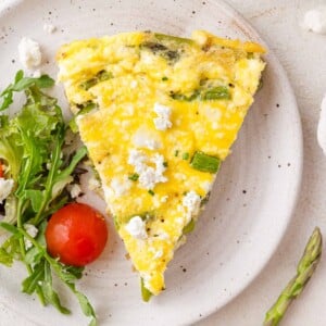 A slice of vegetable frittata with asparagus and goat cheese on a plate, accompanied by mixed greens and a cherry tomato.