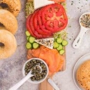 Bagel and lox board with smoked salmon, tomatoes, onions, alfalfa sprouts, cucumber slices, cream cheese, capers, and seasonings, accompanied by bagels.