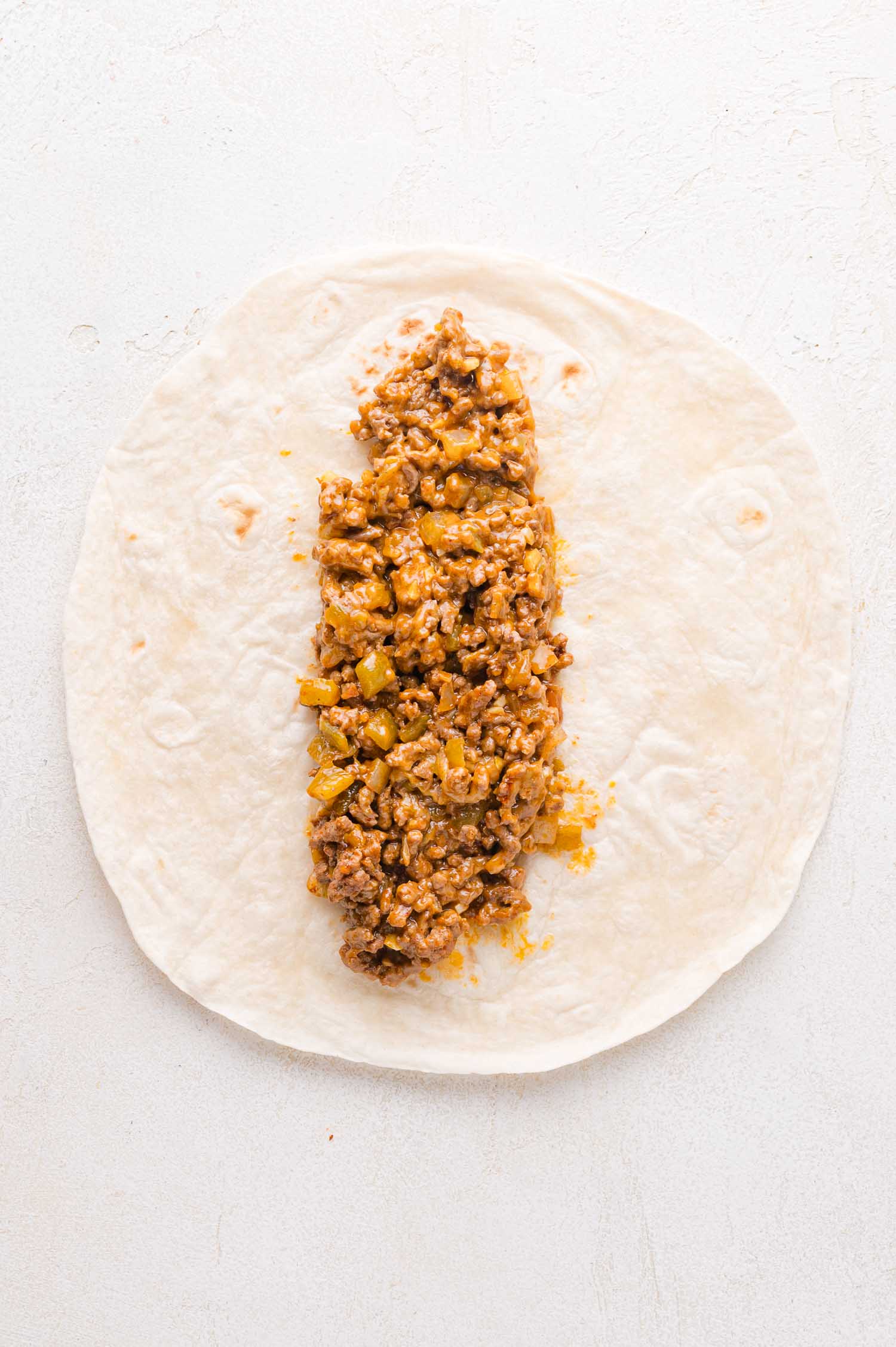 Uncooked enchilada on a light surface with a filling of ground meat, diced green peppers, and melted cheese placed in the center of a flour tortilla.