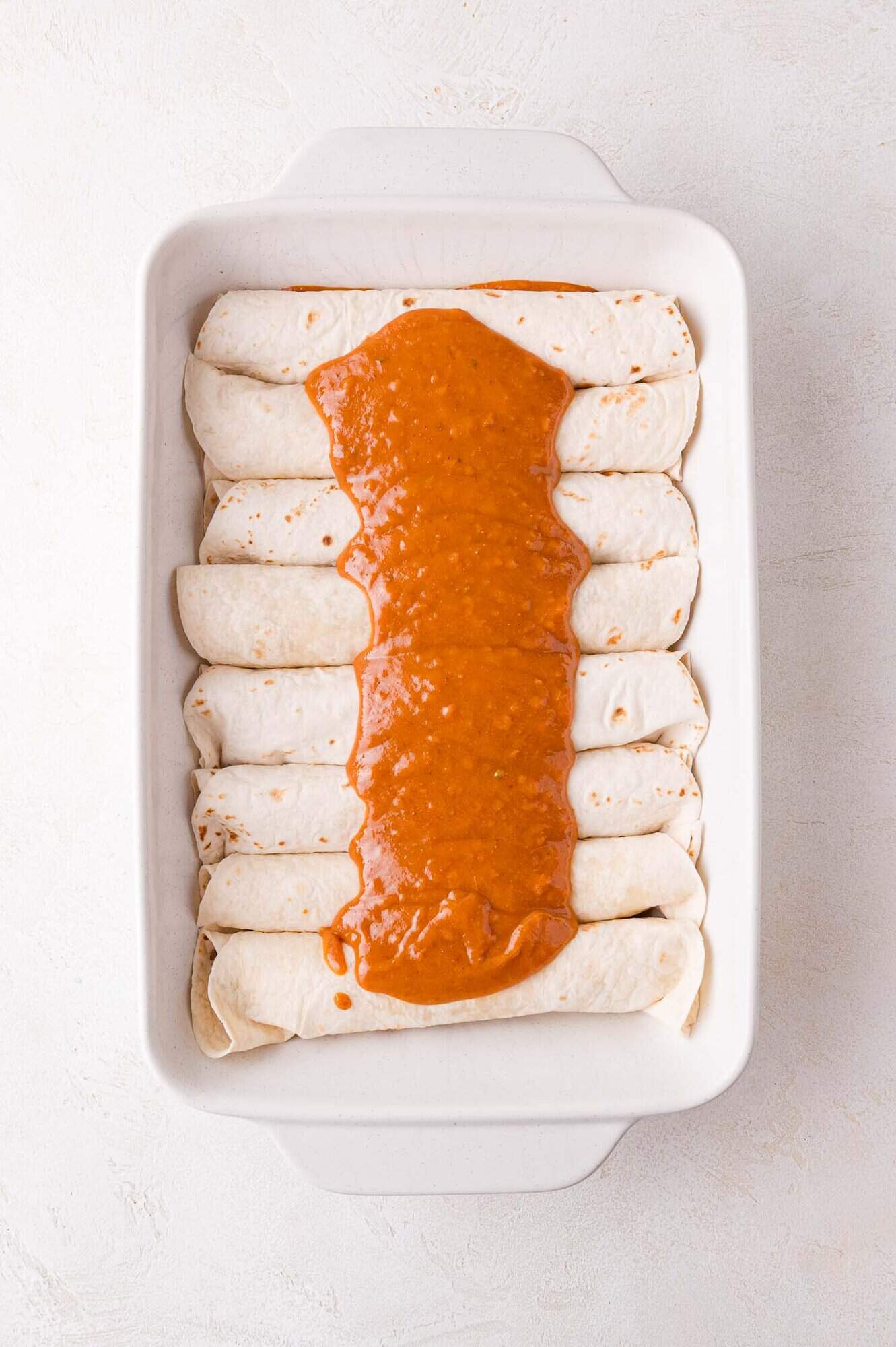 A baking dish with evenly arranged rolled tortillas topped with a thick layer of red sauce down the middle.