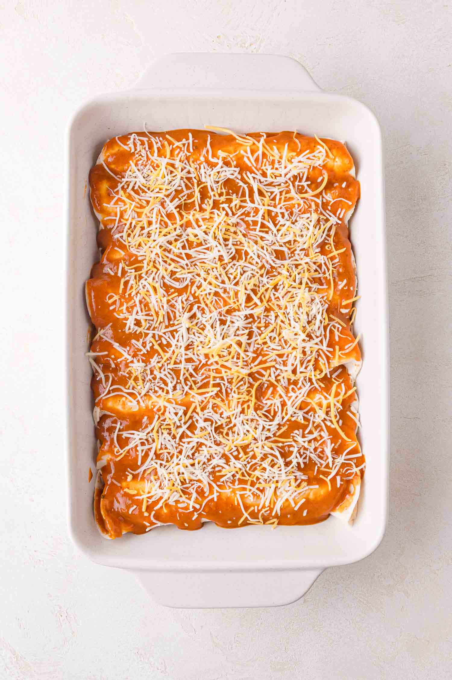 A white baking dish filled with enchiladas, topped with orange sauce and shredded cheese, set on a light textured surface.